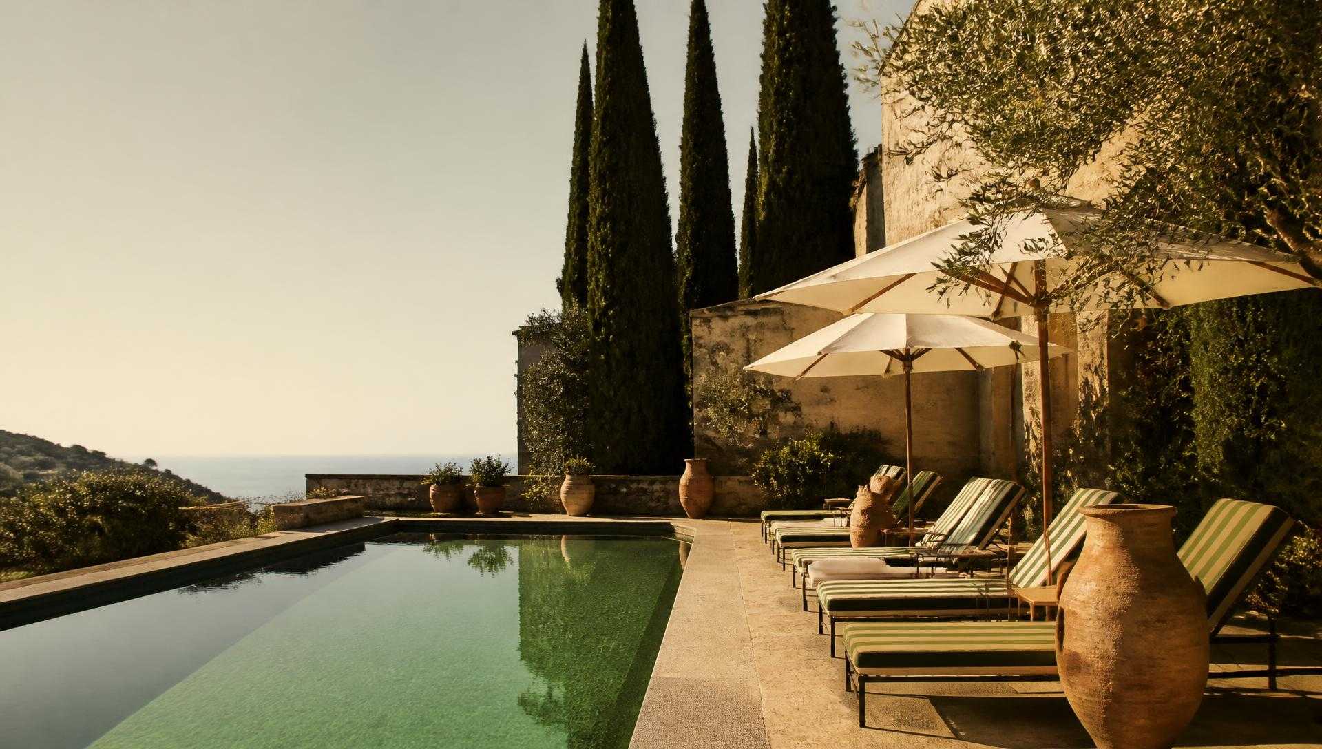 A quiet Mediterranean hotel pool terrace at golden hour, cypress trees and cream umbrellas beside still green water